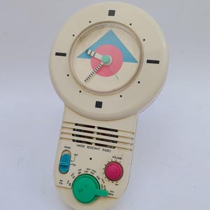 Puede incluir: Radio vintage resistente al agua de color crema con esfera de reloj. El reloj tiene un diseño de círculo rosa y triángulo azul. Cuenta con una perilla de sintonización verde, un dial de volumen rosa y un selector de banda azul. La radio tiene ajustes AM/FM.