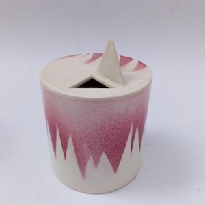 Può includere: Un contenitore cilindrico in ceramica con base color crema e parte superiore e corpo rosa maculato. Il corpo presenta un motivo frastagliato, simile a una montagna, in rosa. Un picco triangolare si erge dalla parte superiore, con un'apertura triangolare sottostante.