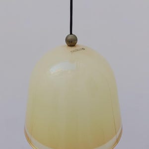 May include: A pendant light fixture with a cream-colored, bell-shaped glass shade. The shade is suspended by a black cord and a small, round, brass-colored fixture. The light fixture has a minimalist design.