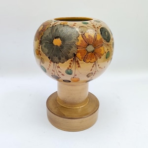 Peut inclure: Un vase en céramique avec un dessus rond et une base en piédestal. Le vase est décoré de grandes fleurs stylisées dans des tons de marron, d'orange et de vert sur un fond beige. La base est de couleur marron clair.