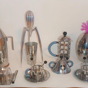 Könnte beinhalten: Eine Sammlung silberner Küchenutensilien, darunter ein Zitruspresse, eine Kaffeemaschine, eine Teekanne, ein Zuckerstreuer und ein Milchkännchen. Die Utensilien sind auf einer weißen Oberfläche angeordnet.