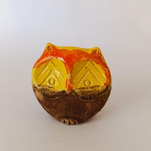 May include: A ceramic owl figurine with a yellow body, orange accents, and brown base. The owl has detailed eyes and a textured surface, showcasing a handcrafted aesthetic. This decorative item is suitable for home decor.