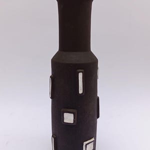 Pode incluir: Um vaso de cerâmica castanho escuro com corpo cilíndrico e topo alargado. O vaso apresenta detalhes retangulares e quadrados prateados. O vaso tem aproximadamente 30 cm de altura, adequado para exibir flores ou como peça decorativa.