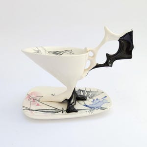 May include: A unique white ceramic teacup with a black and white handle and a matching saucer. The cup has a triangular shape and is decorated with black line drawings. The saucer features pink and blue watercolor designs.