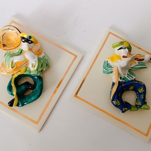 Può includere: Due figurine in ceramica di musicisti, una che suona la tromba e l'altra il violino. Entrambe le figurine sono dipinte con colori vivaci, tra cui verde, giallo, blu e bianco. Le figurine sono montate su piastrelle quadrate con un bordo dorato.