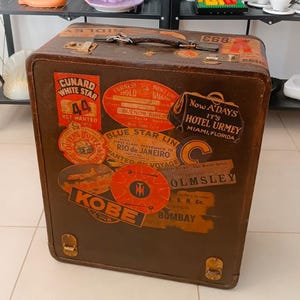 Op de afbeelding: Een vintage bruine leren koffer met een versleten, verweerde uitstraling. Hij is bedekt met kleurrijke reisstickers van verschillende bestemmingen, waaronder "CUNARD WHITE STAR", "BLUE STAR LINE", "KOBE", "OLMSLEY" en "NOW A'DAYS IT'S HOTEL URMEY MIAMI, FLORIDA".