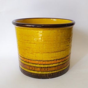 Op de afbeelding: Een gele keramische plantenpot met een bruine rand en een geweven patroon. De pot heeft een band met bruine, oranje en gele strepen rond het midden.