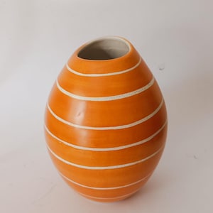 Pode incluir: Vaso de cerâmica laranja com listras horizontais brancas. O vaso tem uma forma arredondada, em forma de ovo, com uma abertura larga no topo. O interior é cinza claro.