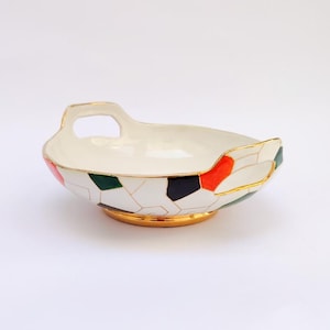 Può includere: Una ciotola decorativa in ceramica con interno ed esterno bianchi, con un motivo geometrico di forme rosse, verdi e nere delineate in oro. La ciotola ha un bordo e una base dorati e un manico su ogni lato.
