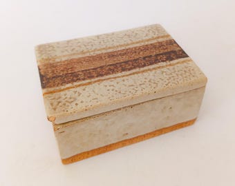 May include: A rectangular, beige ceramic box with a lid. The lid features three horizontal brown stripes. The box has a textured surface and a thin orange stripe along the bottom edge. The box is closed.