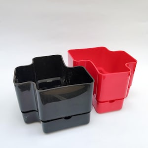 以下が含まれることがあります： 2つのプラスチック製のデスクオーガナイザー。1つは黒、もう1つは赤です。どちらも、複数のコンパートメントとベースに小さな引き出しを備えた、ユニークな波型のデザインです。オーガナイザーは白い背景に配置されています。