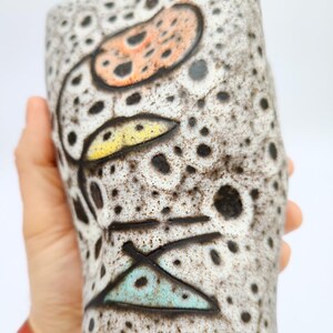 Puede incluir: Un jarr&oacute;n de cer&aacute;mica con una superficie moteada y texturizada. El jarr&oacute;n presenta un dise&ntilde;o floral con acentos naranja, amarillo y turquesa. Puntos y l&iacute;neas negras se suman al dise&ntilde;o art&iacute;stico. El jarr&oacute;n se sostiene en una mano.