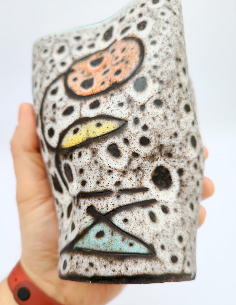 Puede incluir: Un jarr&oacute;n de cer&aacute;mica con una superficie moteada y texturizada. El jarr&oacute;n presenta un dise&ntilde;o floral con acentos naranja, amarillo y turquesa. Puntos y l&iacute;neas negras se suman al dise&ntilde;o art&iacute;stico. El jarr&oacute;n se sostiene en una mano.