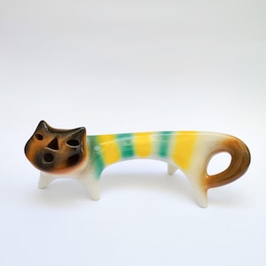 May include: A ceramic cat ornament with a brown and tan face, white legs, and a striped body in yellow and green. The tail has a brown loop. The cat is standing on a white surface.