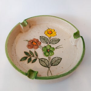 May include: A white ceramic ashtray with a green rim and a floral design. The flowers are yellow, orange, brown, and green.