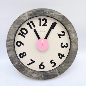 Pode incluir: Um relógio de parede redondo com uma moldura de padrão de mármore cinzento e um mostrador branco sujo. O relógio apresenta grandes numerais pretos de 1 a 12, ponteiros pretos de horas e minutos e um detalhe circular rosa central.