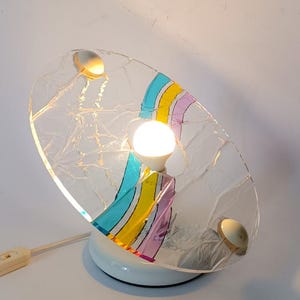 May include: A white table lamp with a clear glass shade featuring a colorful rainbow stripe design. The lamp has a white base and a cord with an on/off switch.