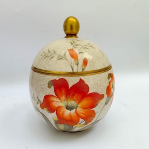Puede incluir: Un tarro decorativo redondo con tapa, fondo crema y diseño floral. El tarro presenta una gran flor naranja vibrante y capullos naranjas más pequeños. La tapa y la base están ribeteadas en dorado, y el remate es dorado.