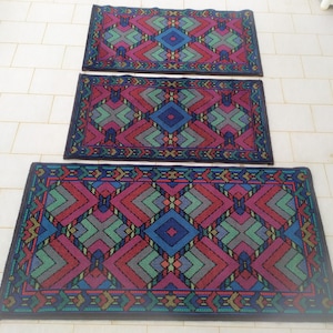 May include: Three colorful patterned rugs with geometric designs in shades of red, blue, green, and gray. The rugs are arranged in a vertical stack, with the largest rug at the bottom and the smallest rug at the top.