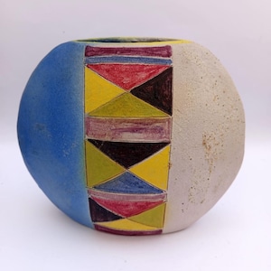 May include: A ceramic vase with a rounded shape, featuring a color-blocked design. One side is a solid blue, the other is a textured off-white. The center panel has a geometric pattern of triangles and rectangles in yellow, red, and black.