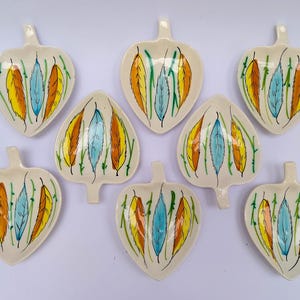 Puede incluir: Ocho platos de cerámica con forma de hoja de color crema. Cada plato presenta un diseño pintado a mano de plumas coloridas en amarillo, naranja y azul, con tallos verdes. Los platos tienen un pequeño asa en la parte superior.