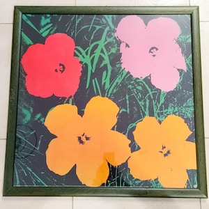 May include: A framed print featuring four large, stylized flowers in vibrant colors. The flowers are red, pink, and orange, set against a dark background with green grass-like strokes. The frame is a dark green color.