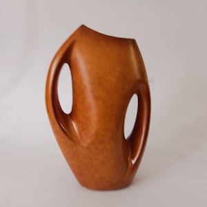 Può includere: Un vaso in ceramica marrone dalla forma organica unica. Il vaso presenta due grandi manici aperti e una superficie strutturata. La parte superiore ha un design asimmetrico. Il vaso è di un caldo tono terroso, adatto all'arredamento della casa.