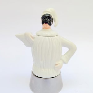 Pode incluir: Uma cafeteira de cerâmica branca em forma de figura com máscara preta, chapéu branco e gola com babados. A base é de metal prateado. A figura tem um rosto pintado e está em pose com um braço levantado.