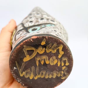 Puede incluir: Un jarr&oacute;n de cer&aacute;mica hecho a mano con un patr&oacute;n moteado gris y negro, acentuado con detalles turquesa, naranja y amarillo. La base es marr&oacute;n oscuro con letras doradas que dicen "Dier main Vallauris".
