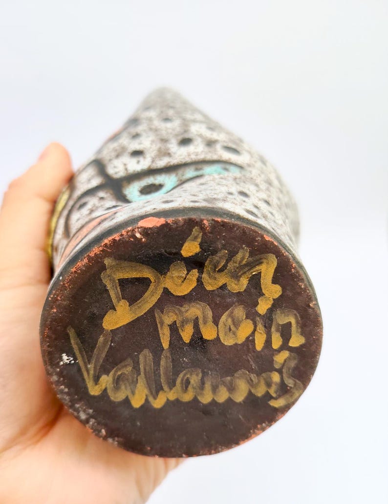Puede incluir: Un jarr&oacute;n de cer&aacute;mica hecho a mano con un patr&oacute;n moteado gris y negro, acentuado con detalles turquesa, naranja y amarillo. La base es marr&oacute;n oscuro con letras doradas que dicen "Dier main Vallauris".