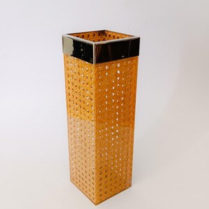 Puede incluir: Un jarrón de mimbre rectangular alto con un borde de metal dorado. El jarrón está hecho de ratán tejido y tiene un color marrón claro.