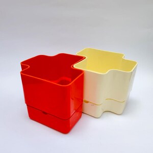 以下が含まれることがあります： 2つのプラスチック容器。1つは鮮やかな赤色、もう1つはオフホワイトです。どちらもユニークな波状の上面と長方形のベースを持っています。容器はおそらく保管または整理用です。
