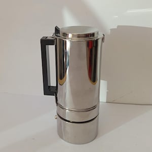 May include: A stainless steel stovetop espresso maker with a black handle. The coffee pot has a cylindrical body with a round base and top. The espresso maker is designed to brew coffee on a stovetop.