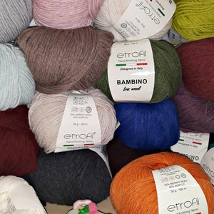 May include: A variety of yarn balls in different colors, including pink, brown, gray, blue, green, and orange. The yarn balls are arranged in a pile on a wooden surface. Each ball has a label with the brand name "etrofil" and the text "Bambino Lux Wool".