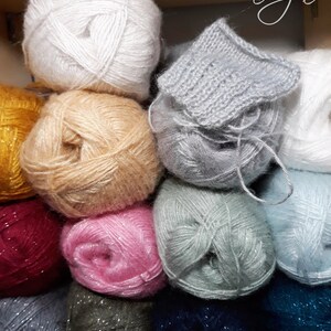 May include: A collection of yarn balls in various colors, including white, yellow, pink, green, blue, and gray. Some of the yarn balls have a sparkly finish. The yarn is arranged in a stack, with the top ball partially covered by a knitted piece of fabric.
