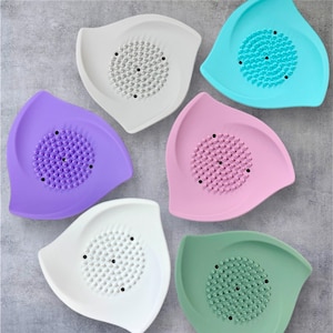 May include: A collection of six leaf-shaped kitchen tools in various colors: white, purple, pink, green, and turquoise. Each tool features a central area with small, raised bumps and a few black dots.