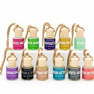May include: A collection of car air fresheners in small glass bottles with wooden caps and hanging cords. Each bottle has a label with a different scent name, such as "Boujee Babe" and "Lavender Sage". The bottles are arranged against a white background.