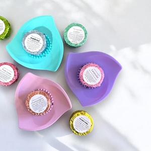 May include: Assortment of six shower steamers in various colors and scents, including lavender, champagne, caramel honey, wanderlust, calm, and fruity loops. Each steamer is displayed on a small, decorative dish.