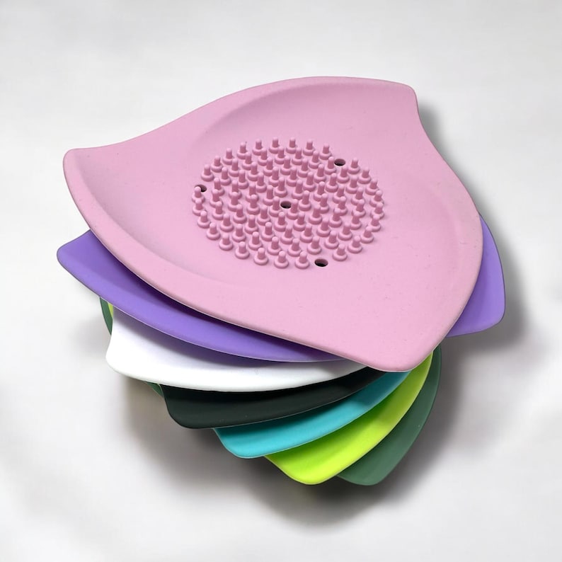 May include: A stack of colorful, leaf-shaped silicone kitchen scrubbers. The top scrubber is pink with a central textured area. Other colors include purple, white, dark green, teal, lime green, and forest green.
