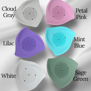 May include: A collection of six leaf-shaped silicone face scrubbers in various colors: cloud gray, petal pink, lilac, mint blue, white, and sage green. Each scrubber features a textured center for exfoliation and the color name is printed on the image.