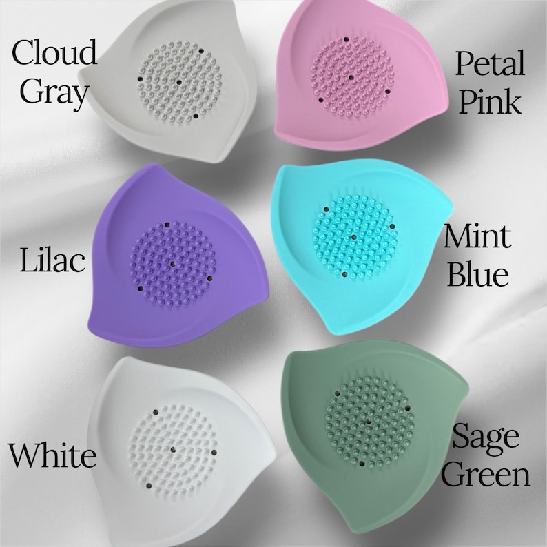 May include: A collection of six leaf-shaped silicone face scrubbers in various colors: cloud gray, petal pink, lilac, mint blue, white, and sage green. Each scrubber features a textured center for exfoliation and the color name is printed on the image.