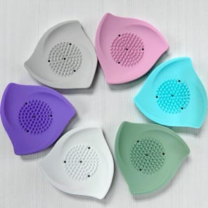 May include: A collection of six colorful, leaf-shaped soap dishes. Each dish features a central, textured area for soap and is in a different pastel color: gray, pink, blue, green, purple, and white. The dishes are arranged in a circular pattern.