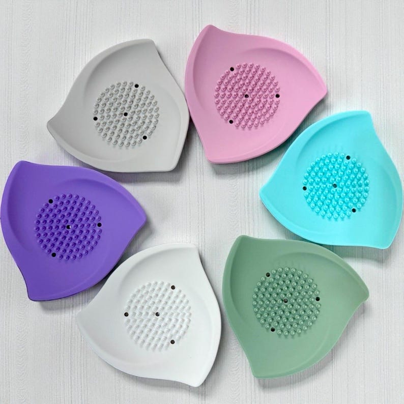 May include: A collection of six colorful, leaf-shaped soap dishes. Each dish features a central, textured area for soap and is in a different pastel color: gray, pink, blue, green, purple, and white. The dishes are arranged in a circular pattern.