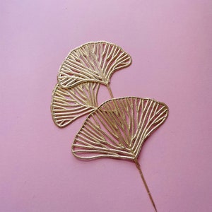 May include: Three gold-coloured ginkgo leaf shapes are attached to a thin gold stem. The leaves are cut out of metal and have a delicate, intricate design. The leaves are arranged in a fan shape, with the largest leaf at the bottom and the smallest leaf at the top.