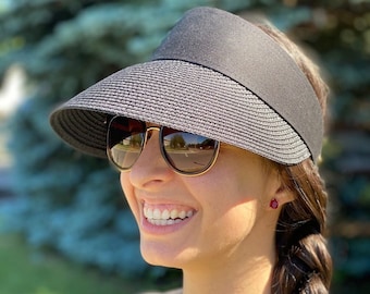 May include: A woman wearing a black visor hat with a wide brim, sunglasses, and a black shirt. She has long brown hair braided down her back and is wearing small red earrings.