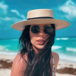 May include: A woman with long black hair wearing a wide-brimmed straw hat with a black band and dark sunglasses. She is standing on a beach with blue water in the background.