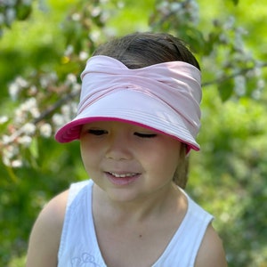 May include: A young person wearing a pink sun hat with a pink and white headband. The hat has a wide brim and a pink underside. The person is wearing a white sleeveless shirt with a floral design.