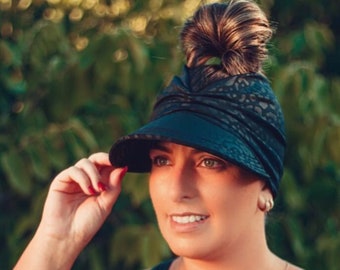 May include: A woman wearing a black one-shoulder swimsuit with a leopard print pattern. She is wearing a black and white patterned hat with a visor. Her hair is pulled back in a bun.