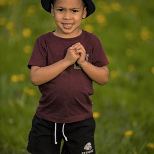 Op de afbeelding: Een jong kind draagt een marineblauwe fedora, een bordeauxrood t-shirt en een zwarte korte broek met een wit trekkoord. De korte broek heeft een witte afbeelding en tekst. Het kind staat in een veld met gele bloemen.