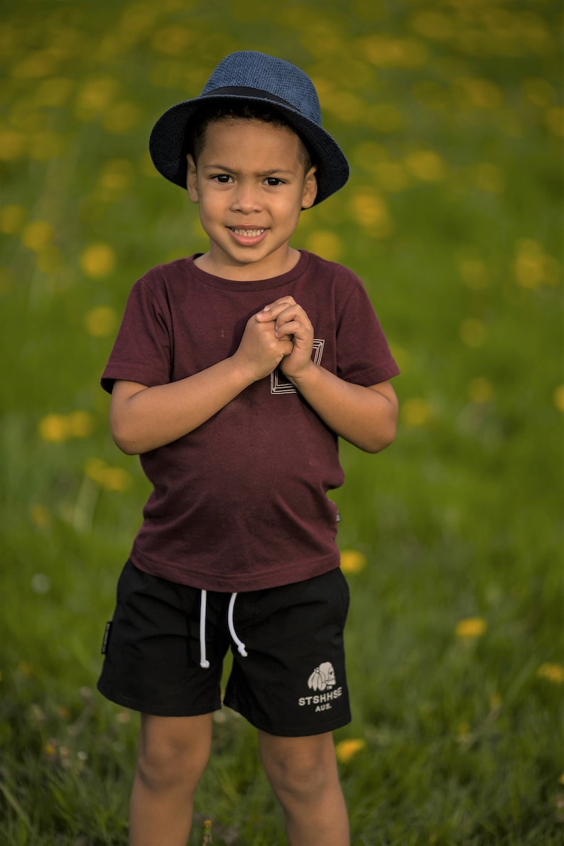 Op de afbeelding: Een jong kind draagt een marineblauwe fedora, een bordeauxrood t-shirt en een zwarte korte broek met een wit trekkoord. De korte broek heeft een witte afbeelding en tekst. Het kind staat in een veld met gele bloemen.